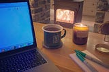 Laptop, mug of tea, candle, sharpies and notebook next to a log burner.