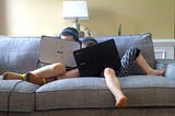 Two kids snuggling on a couch playing on their laptop computers.