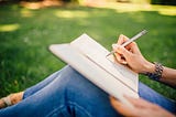 Woman’s hand writing in a notebook, sitting on a grass, wearing blue skinny jeans