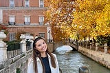A woman wearing a long white coat stands on a bridge overlooking a canal with autum foilage in the background.