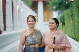 Dos mujeres están de pie y portan vestimenta tradicional tailandesa mientras comparten un abrazo mirando hacia la cámara. Una de ellas usa un bastón.