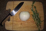 A sharp knife, half of a cut and sliced onion, and two bushy sprigs of fresh rosemary are on a wooden cutting board.