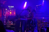 A stage with a Black woman and a keyboard in mics, shrounded in blue.