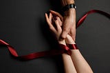 A woman’s hands tied together with red satin ribbon, and held by a man’s hand coming from opposite side of the image, on a dark background