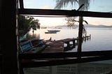 Looking through a window at a ramshackle dock and the sea with small outrigger fishing boats and big bamboo fishing structures scattered around