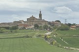 Verdant green fields lead to a town with a church spire rising over all the other buildings. There is a path running through the field and one person is walking towards the town.