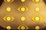 Simple gold handbag with two styles of bright gold “evil” eyes superimposed in a geometric pattern.