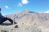 A landscape of a vast grey mountains with the blue sky above, and a girl in a blue jacket sitting in the foreground to the left.