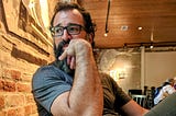 The author with dark curly hair, glasses, and a salt-and-pepper beard sits indoors against an exposed brick wall. He is wearing a grey t-shirt and resting his chin thoughtfully on his hand, looking off to the side. The warm-toned room has a wooden ceiling and worn plaster walls, with other people seated in the background.