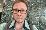 The author has short hair, a beard, and brown framed glasses. He’s wearing a green dress shirt, a grey vest, and is standing in front of a green marble wall. He has a serious face and is looking at the camera.