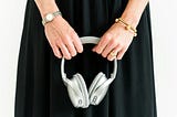 A close-up shot of a mature woman’s hands holding a silver headset. She wears a black dress, a silver watch on one wrist and gold bracelets on the other. She has gold wedding rings on her ring finger.