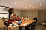 A person giving a presentation to a group of five people sat at a wooden desk