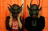 A man and woman hold Mexican devil masks in front of their faces.