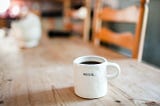 A white coffee mug sits isolated on the table. The coffee nearly reaches the top. One word is on the front in bold black letters. BEGIN.