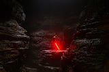 A man with a lightsaber, that's glowing red, in a dark cave. The mood is brooding.