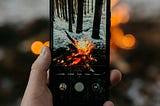 A hand holding a smartphone taking a photo of an outdoor campfire.