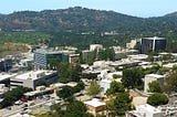 Photograph of JPL from above. A sprawling complex of medium-sized buildings with trees and mountains in the background.