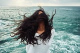 An image of a woman with dark, flowing hair covering her face. The ocean is in the background.
