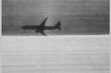 A vintage looking black and white photo of the shadow of an airplane landing. The top have is a dark gray while the bottom half is light gray. A thin gray border surrounded the entire photo.