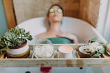 Woman relaxing, leaning her back against the bathhub. The woman is out of focus, there is in front which is in focus. The tray has a candle, plants and crystals in front.