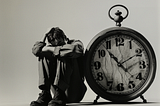 Black and white image of a man sitting on the floor next to a large clock. The man is hugging his knees and stocking his hair in a nervous or stressed emotional state