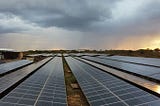 Rows of solar panels in a solar plant with stormy clouds and rain in the distance.