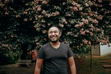 A man in a black shirt smiles in front of a tree.