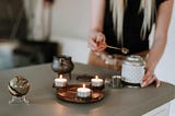 Women standing at a candle lit desk, her adding herbs to a wax scent diffuser appearing to be setting up her spiritual sacred space and altar.