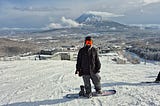 Author standing on a ski slope in snow gear with a snowboard strapped to one foot, you can see Niseko and Mt Yotei in the background