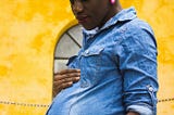 a pregnant femme person with dark brown skin, holding her belly. She wears a blue-jean t-shirt. She has a brightly colored orange, yellow, and green headwrap over a braided updo hairstyle.