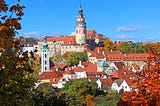 A circular tower, pale pink marble with faded colorful murals around it and a patinaed green bell tower. It stands among a jumble of mostly white buildings with brick-red rooftops. Trees are in full fall colors.