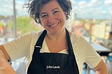 A picture of a smiley Chiara, wearing a white t-shirt and a black apron with the John Lewis brand on it.