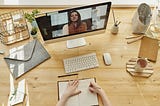 Shot taken from above looking down at a video call on a Mac screen sitting on a wooden surface. Stationary, keyboard, mouse, a book and hands surround the scene.