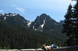 A woman lays on the wall of an overlook in Mt Ranier State Park, mountains, trees, and sky in the background