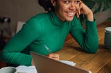 A young woman wearing a green turtleneck leans on a wooden desk with a notepad and laptop. Her face rests against her hand and she has a warm smile.