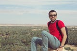 Man sitting in red shirt and jeans and sunglasses on a rock overlooking trees in background