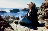 photo of woman smiling in the sun, sitting on a beach with the sea behind