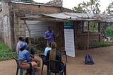 A man stands in front of four people who are sitting in chairs outside a small breezeblock house with a rough corrugated iron verandah. The standing man is speaking and pointing to a display board.