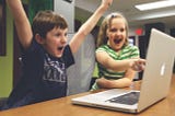 Children win! A young boy and Girl celebrate about what they see on a laptop. Boy puts 2 arms in the air saying Yay! and the girl points excitedly at something on the screen.