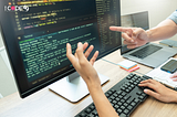 Two developers discussing code on a large monitor in an office setting, with one person pointing at the screen and another gesturing. A keyboard, laptop, smartphone, and notes are visible on the desk. The ICode49 Technolabs logo appears in the top left corner.