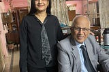 A woman stands next to her father who is seated. Both are indoors in a living room.