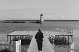 A film photo of a woman in scarf walking towards a pier. We can see a lighthouse in the distance.