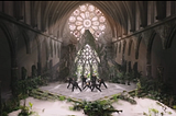 A cathedral with plants growing inside it, 7 dancers in black (BTS) are posed in the center, sunlight streaming through the glass ornamental window in the center back.