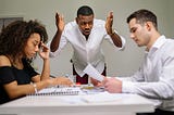 A man angry and gesturing at a table while two people sit and look down at the work that they’ve done, kind of fearful of what that person might say.