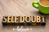 A cup of coffee next to a piece of wood labeled ‘Self Doubt,’ with the word ‘Believe’ written in a graphic style below it