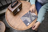 A person sitting at a wooden café table, wearing a green knit sweater, with one hand holding a magazine featuring a bicycle on the cover. A vintage-style portable radio sits on the table.