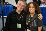 Leo Laporte and Pax sit next to each other and smile for the camera. Both are wearing lanyards with badges. Leo has his arm around Pax, who is wearing an Apple T-shirt.