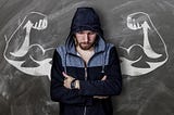 Young white man looking down, and crossing his arms against his chest, wearing a blue and grey hoodie and a serious expression, standing in front of muscular arms drawn on a chalkboard behind him.