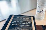 An e-reader in a leather case and a bottle of water. The e-reader’s screen shows the book that the reader is currently reading.