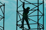 A construction worker stands precariously on scaffolding, suspended in the air, under a blue sky.